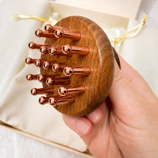 Hand holding the Copper & Sandalwood Scalp Massage Comb close-up, showing all copper prongs, gift box and pouch in the background