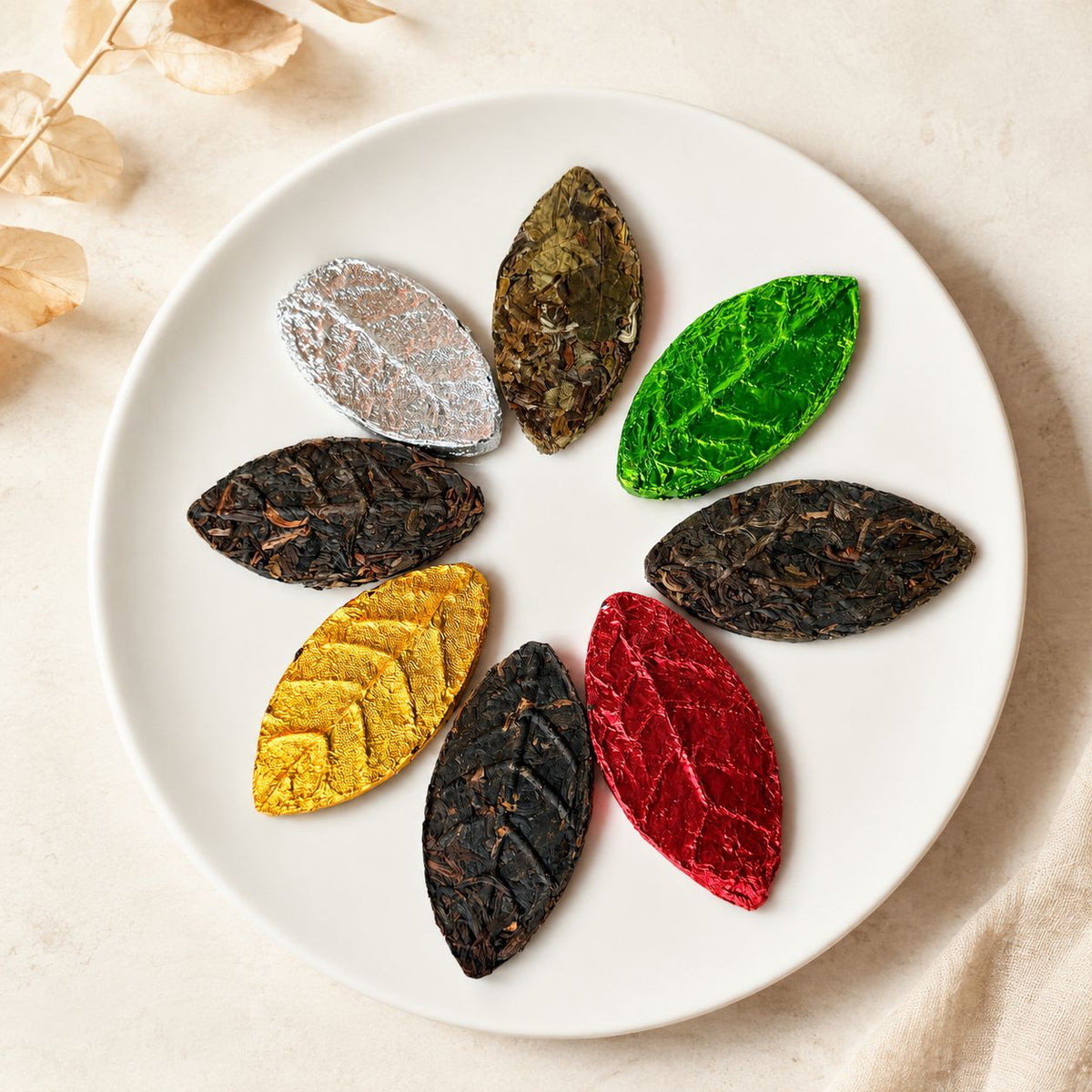 Eight pressed tea leaves in autumn leaf shapes — silver, green, gold and red — arranged like a flower on a white plate