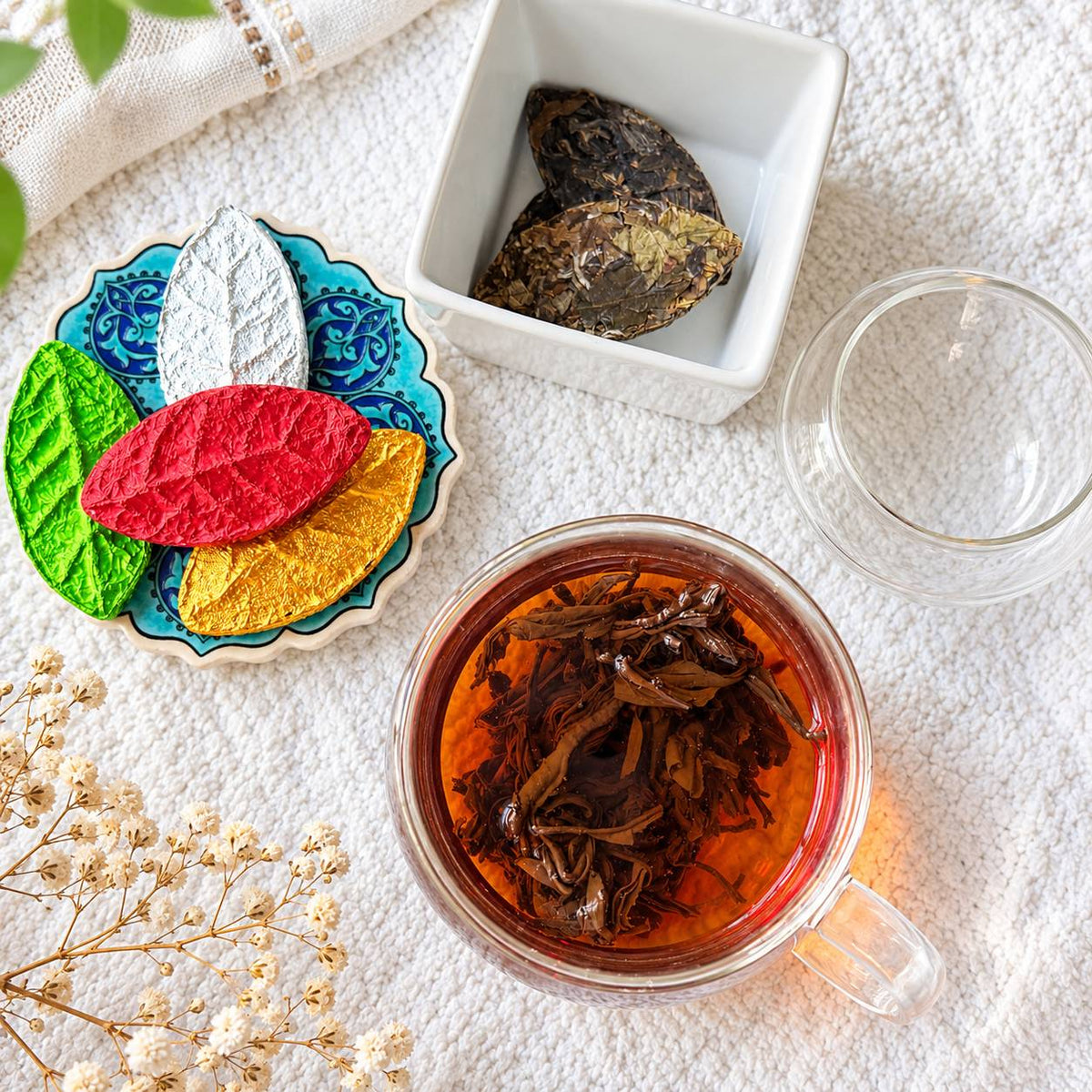 Colorful pressed tea leaves on a blue coaster with a dark pressed leaf in a bowl and a glass of freshly brewed black tea