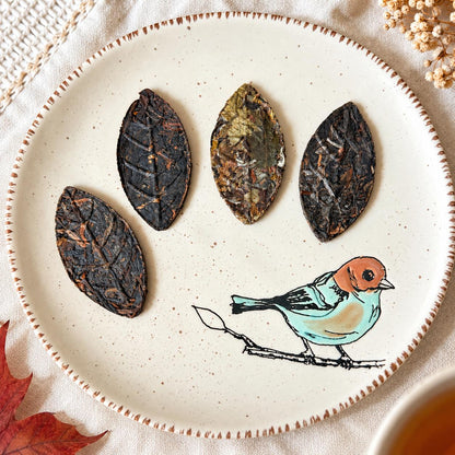 Four dark pressed tea leaves on a speckled plate decorated with a painted bird