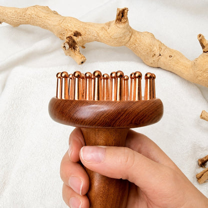 Copper & Rosewood Deep Tissue Massager held by the handle, copper teeth facing up, driftwood behind