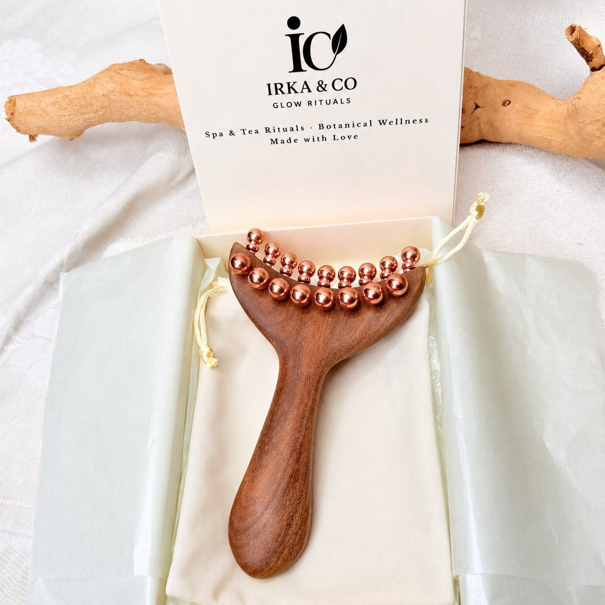 Copper & Wood Deep Tissue Massage Tool in its open IRKA & CO gift box with the Glow Rituals branded insert card, driftwood in the background