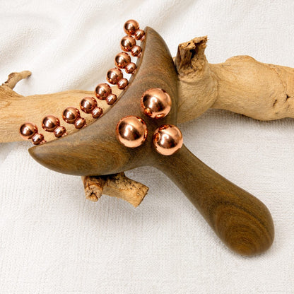 Angled artistic shot of the Copper & Wood Deep Tissue Massage Tool resting on driftwood, showing the curved bead row and three larger copper balls