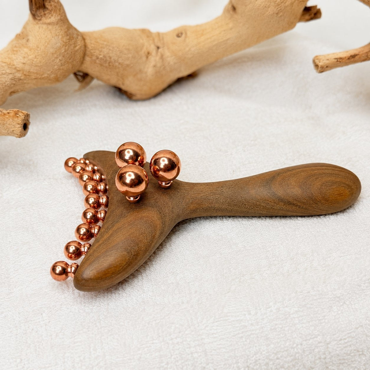 Side-angle view of the Copper & Wood Deep Tissue Massage Tool lying on textured white cloth in front of a driftwood branch