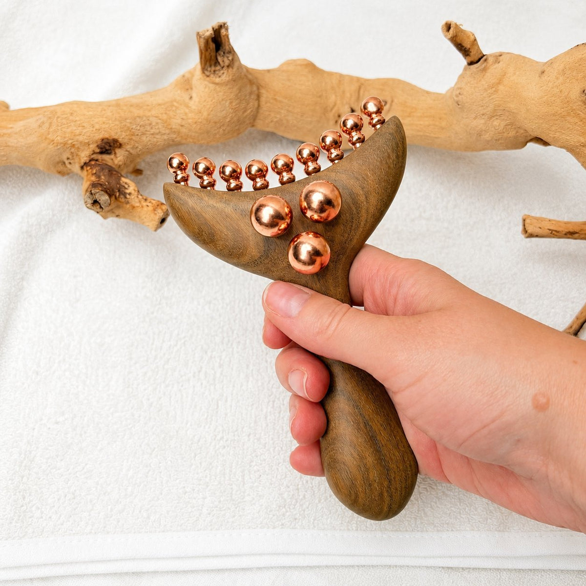 Hand holding the Copper & Wood Deep Tissue Massage Tool upright by its ergonomic handle against a driftwood and cloth backdrop