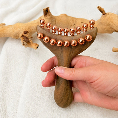 Front-facing close-up of the Copper & Wood Deep Tissue Massage Tool held in hand, showing the full double row of copper massage balls along the curved head