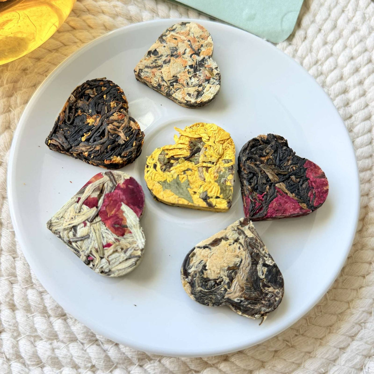 Seven heart shaped compressed tea hearts on a white plate — assorted black, white and pu-erh teas with visible rose petals, chrysanthemum and osmanthus flowers