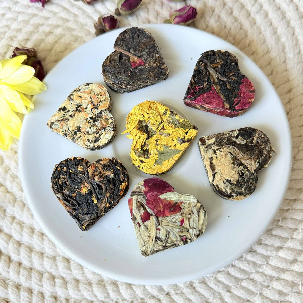 Heart shaped tea sampler arranged on a white plate with a yellow flower and dried rose buds on a cream woven background