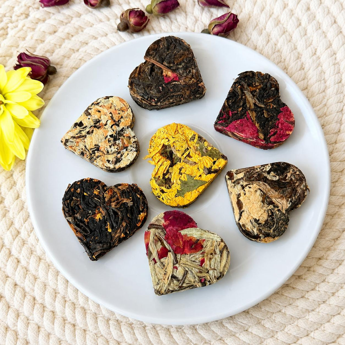 Seven assorted heart shaped compressed tea hearts arranged on a white plate with dried rose buds and a yellow flower