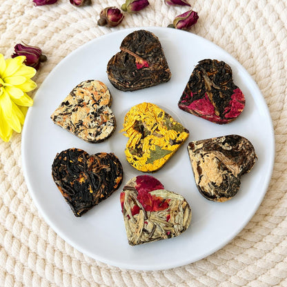 Seven assorted heart shaped compressed tea hearts arranged on a white plate with dried rose buds and a yellow flower