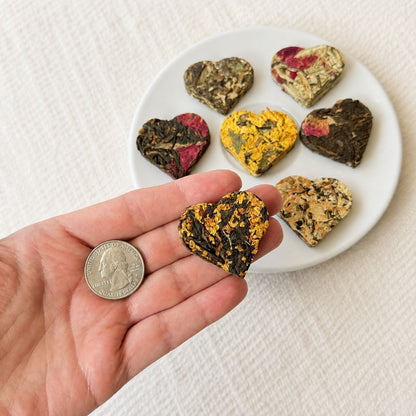 Heart shaped tea heart held in a hand next to a quarter coin for scale, with the tea sampler plate behind