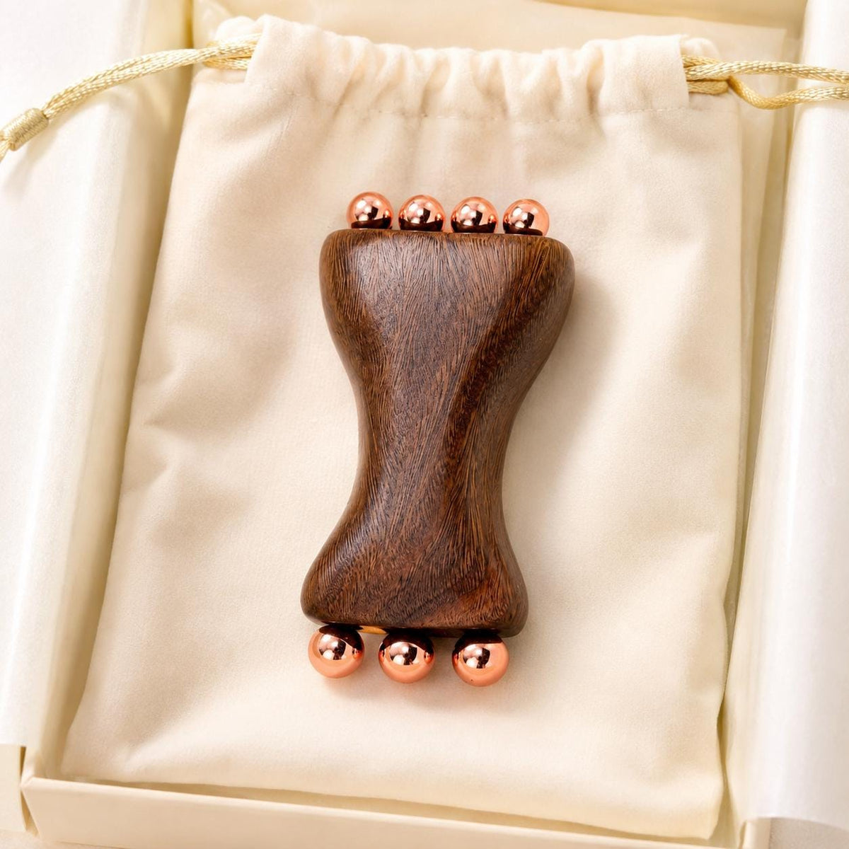 Copper & Sandalwood Facial Massage Tool resting on a cream drawstring pouch inside its gift box, top-down view showing both rows of copper massage balls