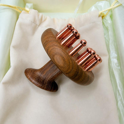 Copper & Sandalwood Scalp Massage Comb resting on a cream drawstring pouch, side angle showing the round wooden head and copper prong teeth