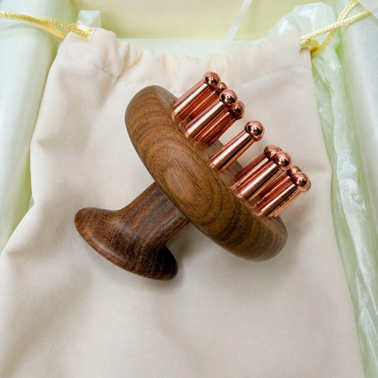 Copper & Sandalwood Scalp Massage Comb resting on a cream drawstring pouch, side angle showing the round wooden head and copper prong teeth