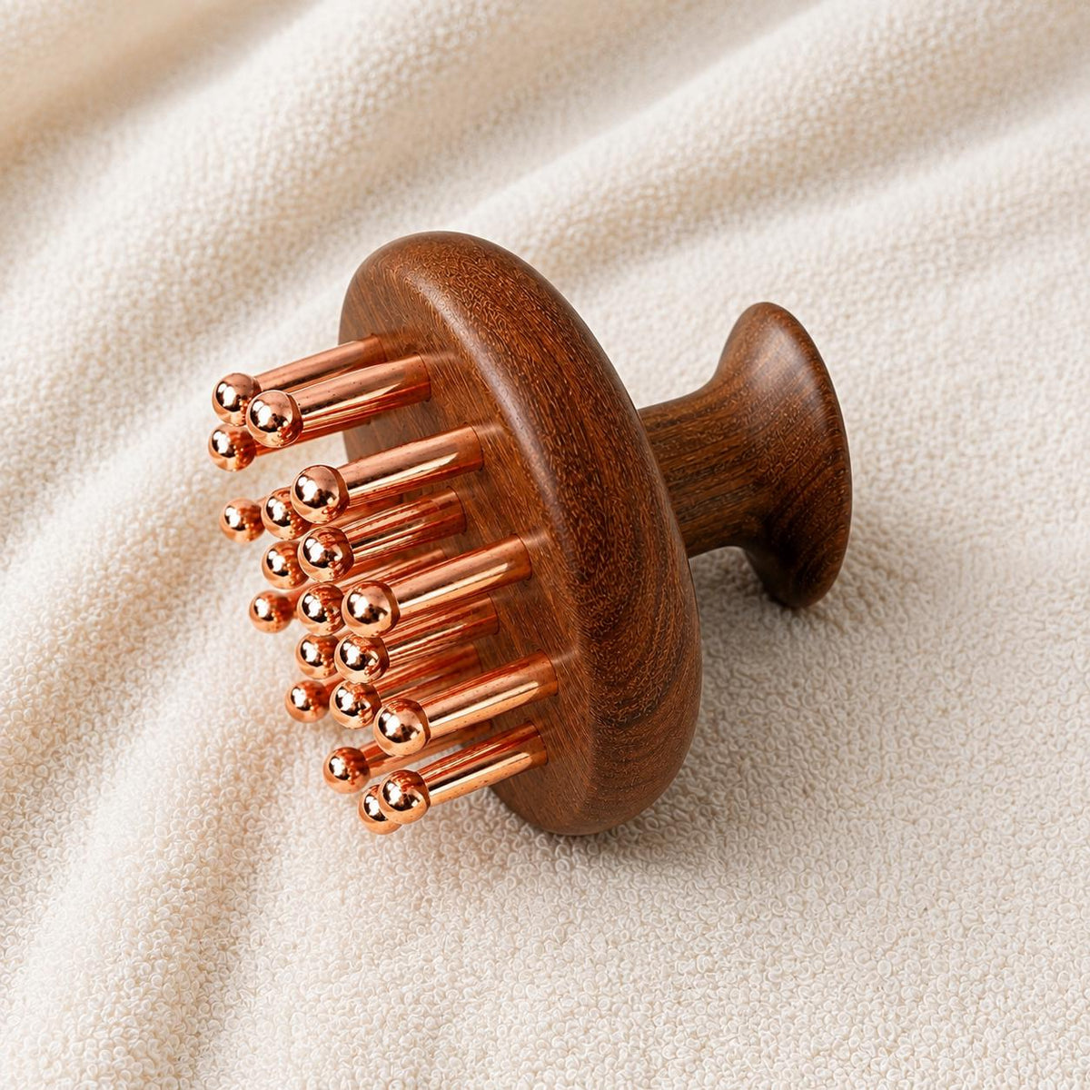 Copper & Sandalwood Scalp Massage Comb on a textured cream towel, angled view showing the round wooden head with copper prongs and mushroom-shaped handle