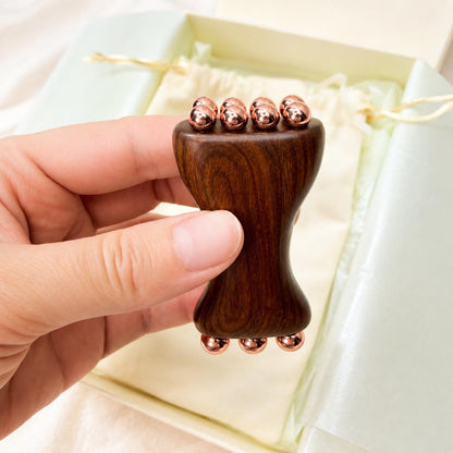 Hand holding the Copper & Sandalwood Facial Massage Tool, its hourglass wooden shape with copper roller balls on both ends, over the open gift box